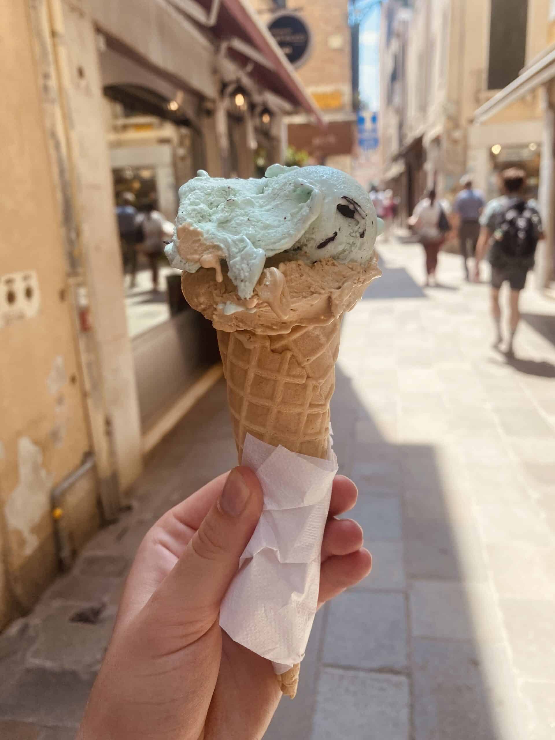 Hand holding an ice cream cone with caramel and mint chip scoops in Venice, Italy, with a cobblestone walkway in the background — food category cover image.